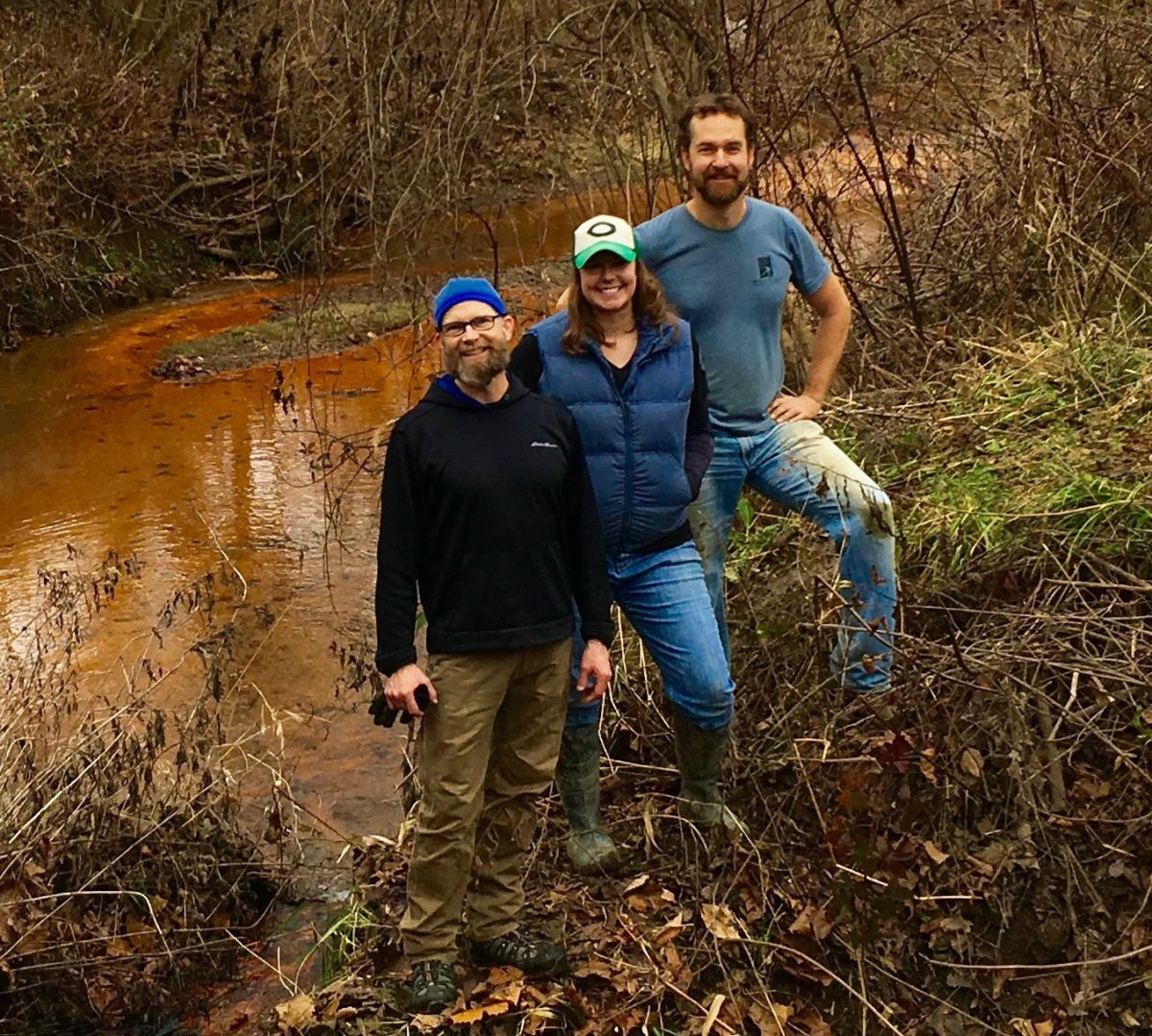 The 3-person reclaimed team standing in front of a rusty river